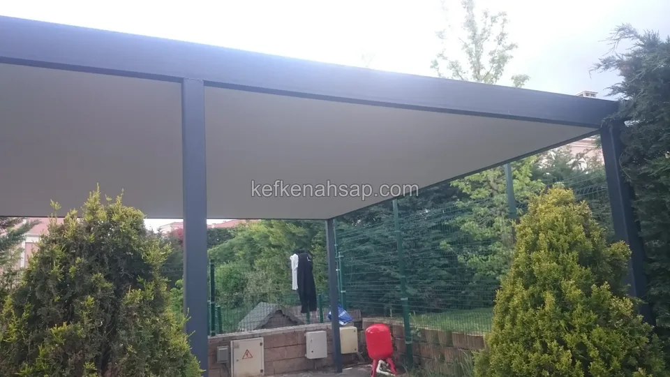 Antrasit metal direkli geniş pergola, üstü beyaz örtülü, önde sarı yeşil çalılar ve arkada bahçe çiti