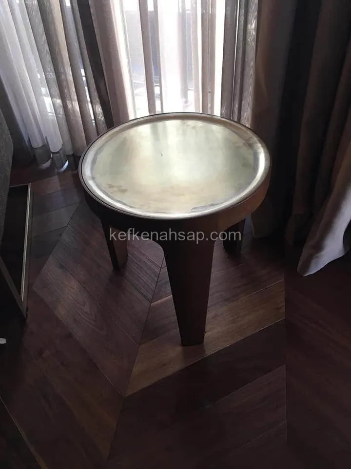 Koyu ahşap gövdeli, üstü pirinç renkli metal tepsi tablalı yuvarlak yan sehpa, balık sırtı parke zemin üzerinde duruyor