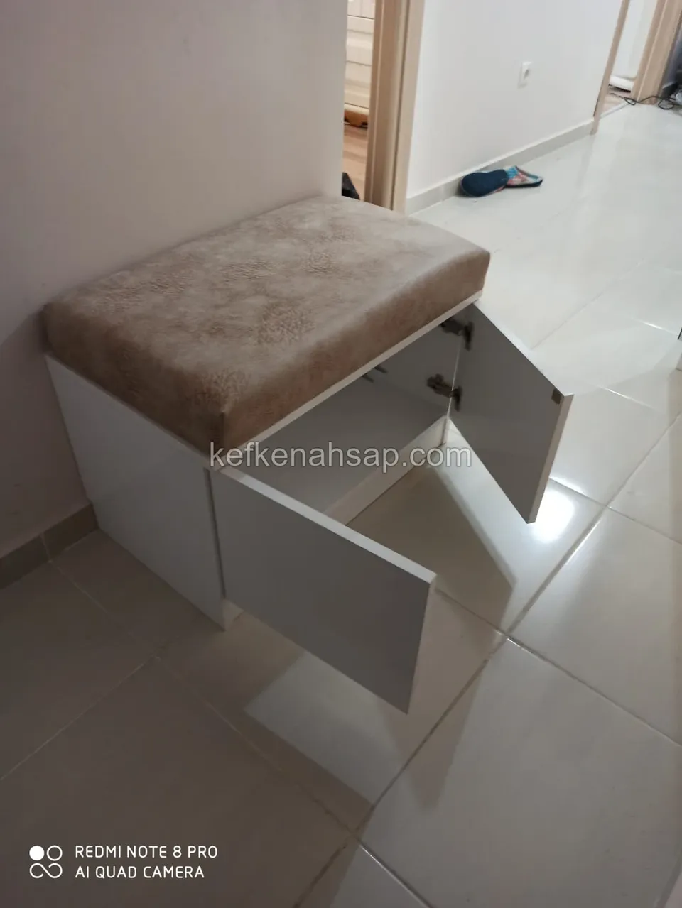 White storage bench with beige padded seat and open doors in a hallway with light ceramic tile floor
