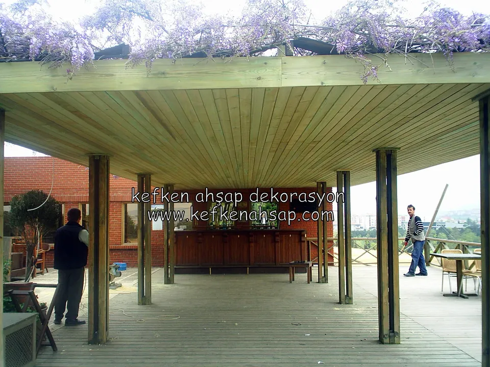 Üstü mor salkımlarla kaplı geniş pergola çatısı, altı lambri kaplı, ahşap deck ve kenarda iki kişi