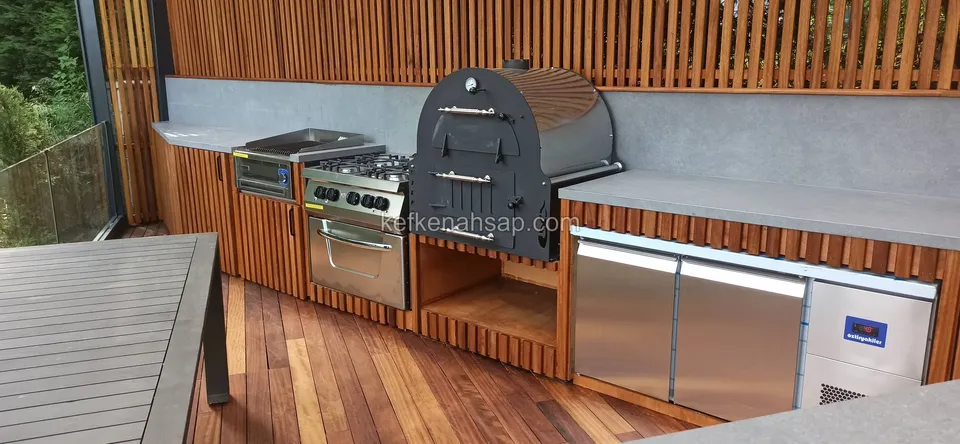 Außenküche mit Holzbackofen, Herd und Edelstahlkühlschrank vor einer Lattenwand, links eine Tischecke