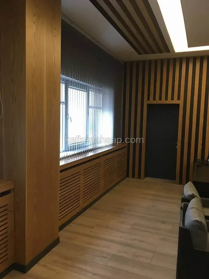 Ofis odasında ahşap lamba kaplı duvar ve tavan, pencere altında ızgaralı ahşap kalorifer kapağı, koyu renk kapı