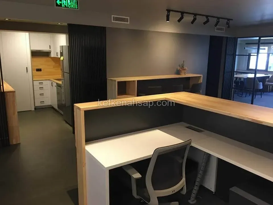 Açık renk masif ahşap üstlü L şekilli ofis bankosu, arkada beyaz mutfak dolapları, siyah çıtalı panel ve cam bölme