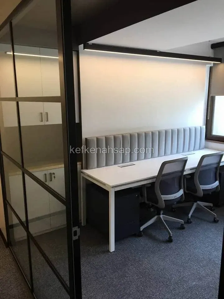 Siyah çerçeveli cam bölmeli ofiste beyaz uzun çalışma masası, gri dikey kapitone duvar paneli ve iki ofis koltuğu