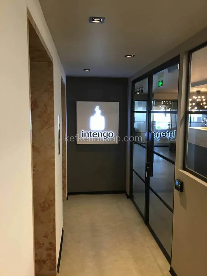 Siyah profilli cam kapı ve aydınlatmalı tabela bulunan modern ofis giriş koridoru.