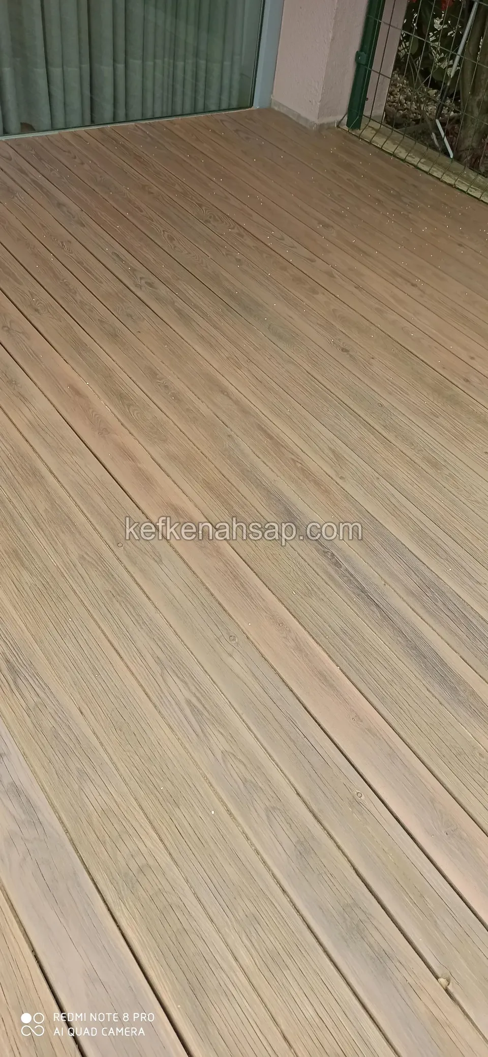 Nahaufnahme graubrauner geriffelter Terrassendielen, oben Glastür und Vorhang