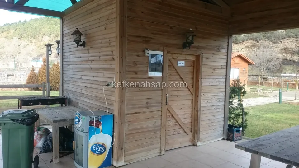 Üstü örtülü terasta tahta duvar kaplaması ve çapraz çıtalı ahşap kapı; önünde masalar, yanda çöp kovası ve duvar lambaları