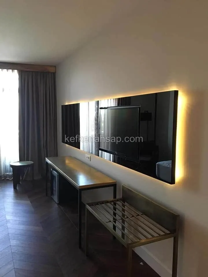 Otel odasında duvara monte arkadan aydınlatmalı koyu panel, TV, uzun çalışma masası, bavul sehpası ve balık sırtı parke zemin