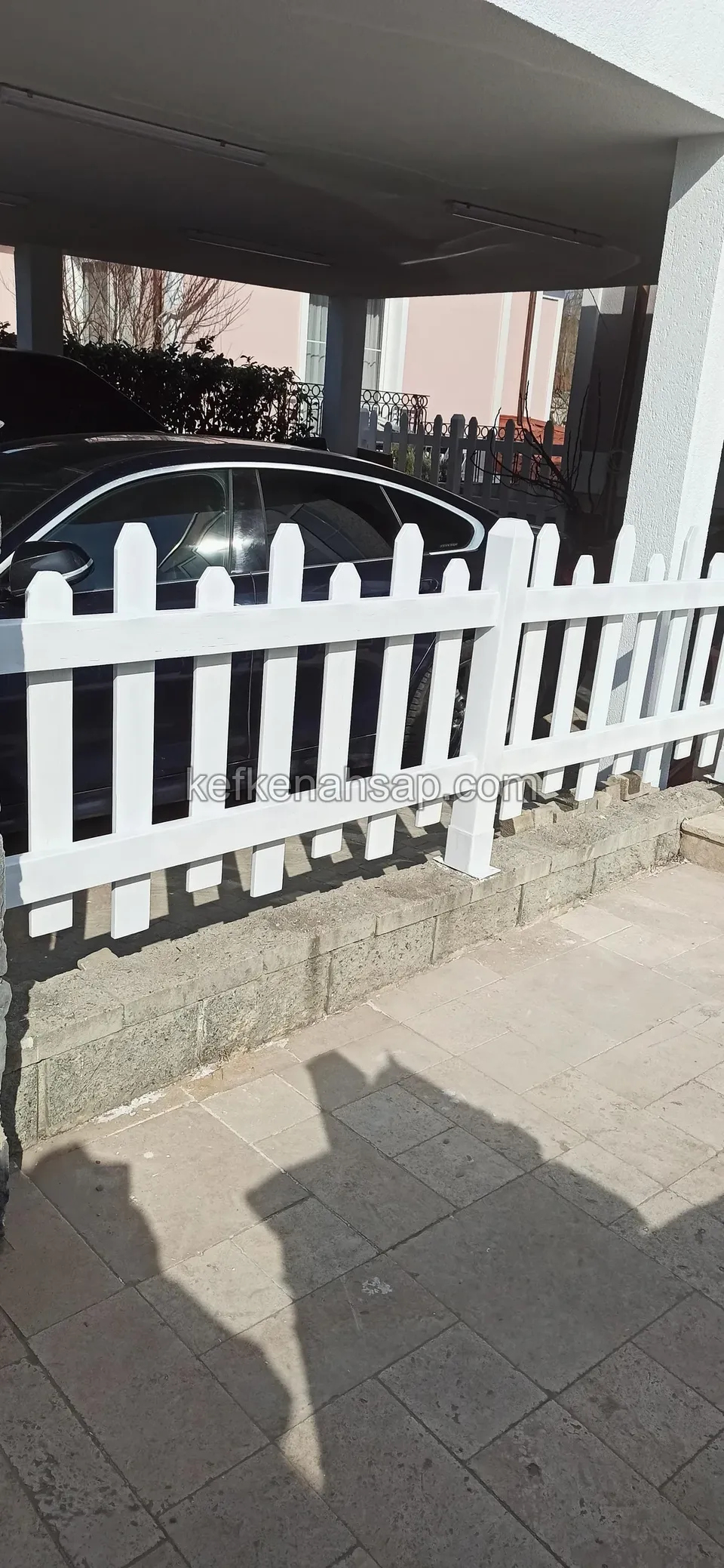 Taş duvarın üstünde uzanan beyaz boyalı çıtalı çit, arkada kapalı otopark ve araç