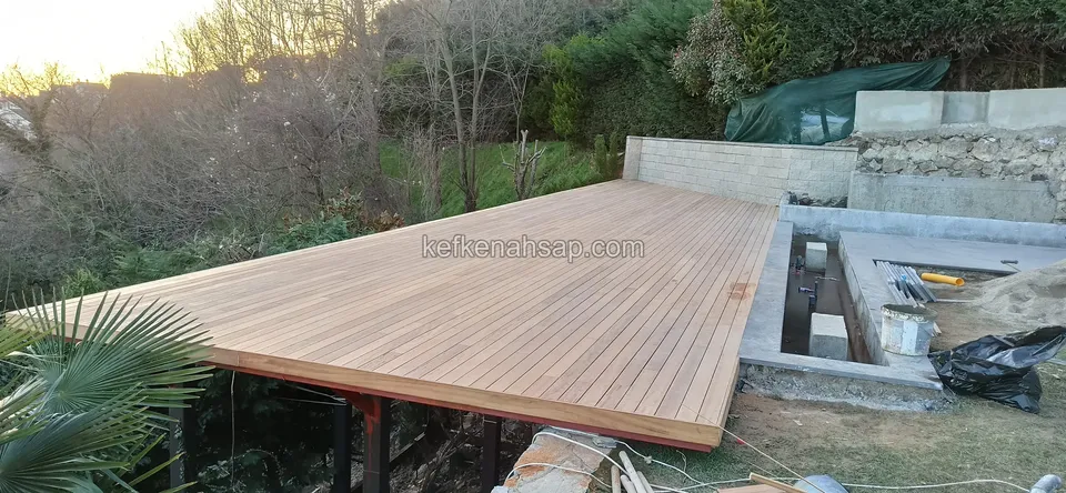 Palmiye ve yamaç önünde uzanan uzun, açık renkli yükseltilmiş ahşap deck platformu