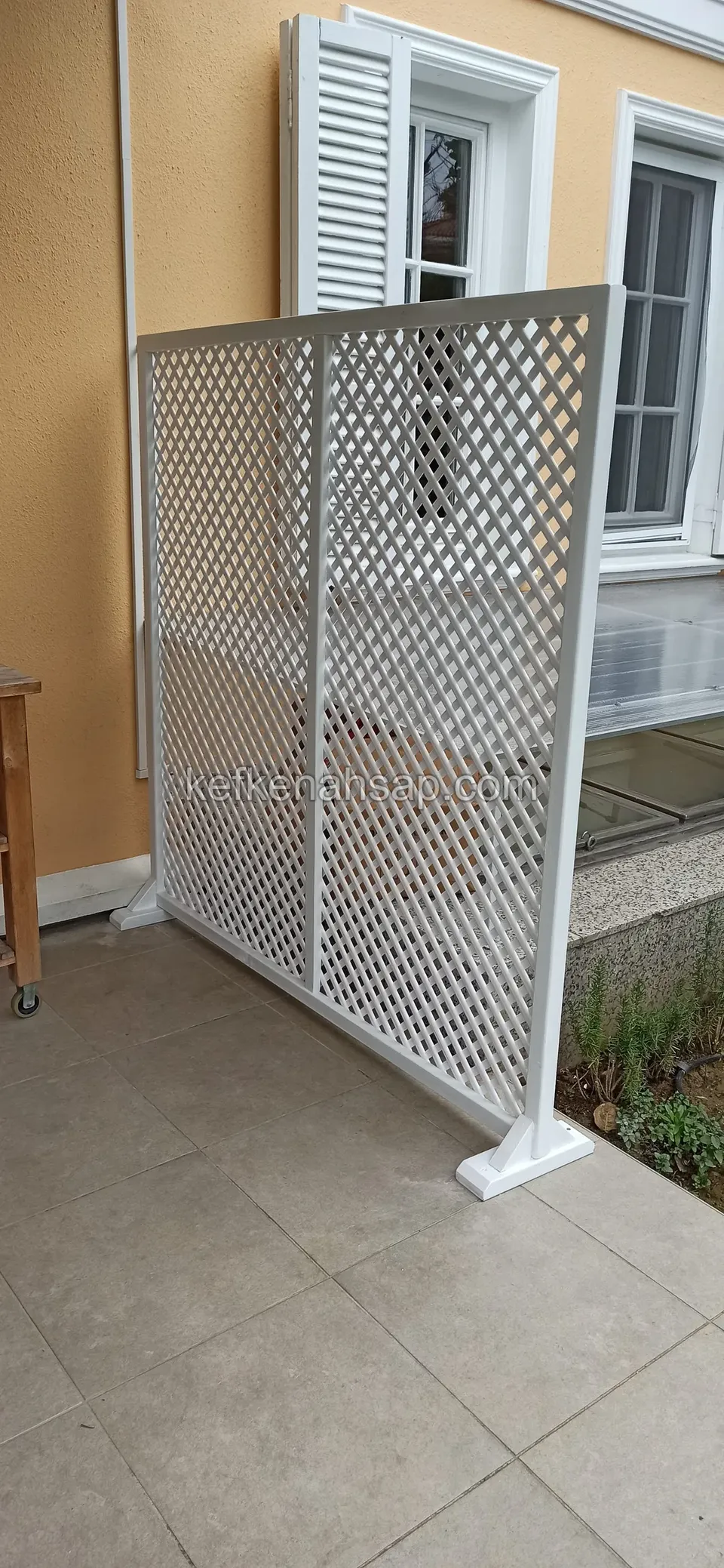 Panjurlu pencerenin ve camlı kapının yanında, terasta duran beyaz boyalı çaprazlı kafes panel; yan taraftan görünüş