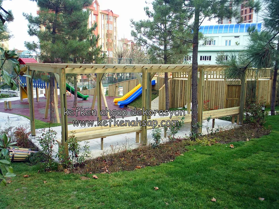 Çocuk oyun alanının önünde, çimenlik içinde ahşap pergola, uzun bank ve yanında açık tonlu ahşap çit