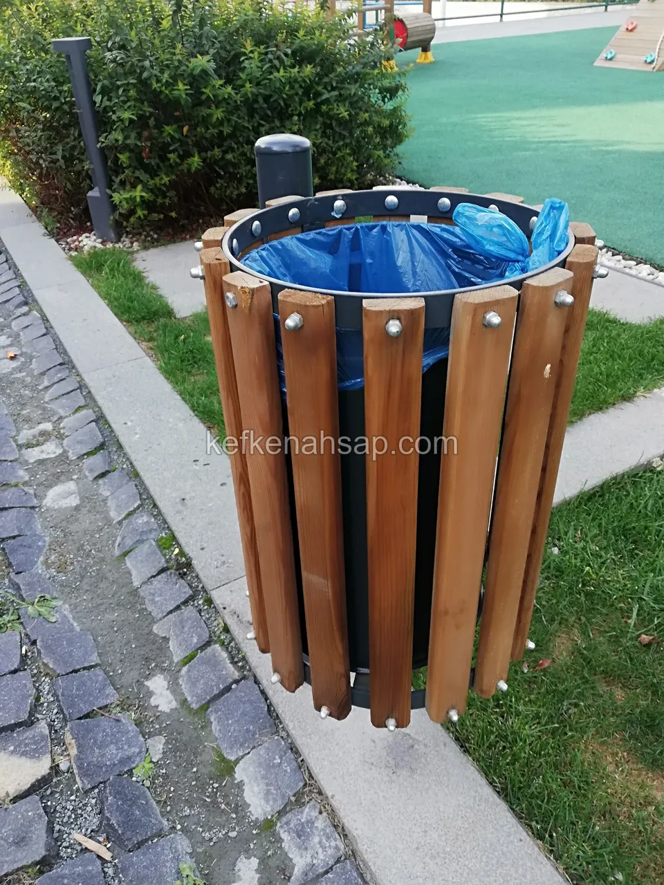 Parkta çimen ve parke taş yolu kenarında, çevresi dikey ahşap çıtalarla kaplı metal çöp kovası, içinde mavi poşet