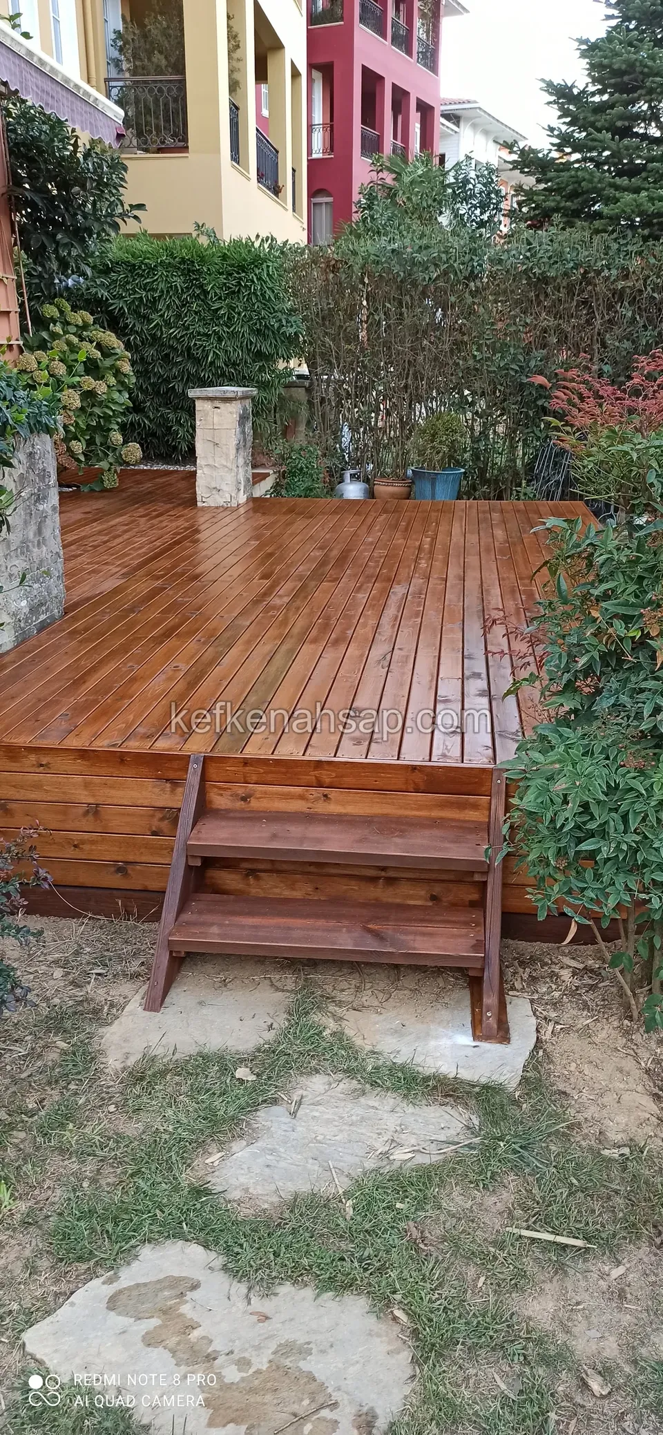Glossy varnished deck with a timber step, stone stepping slabs in front and a hedge and house behind