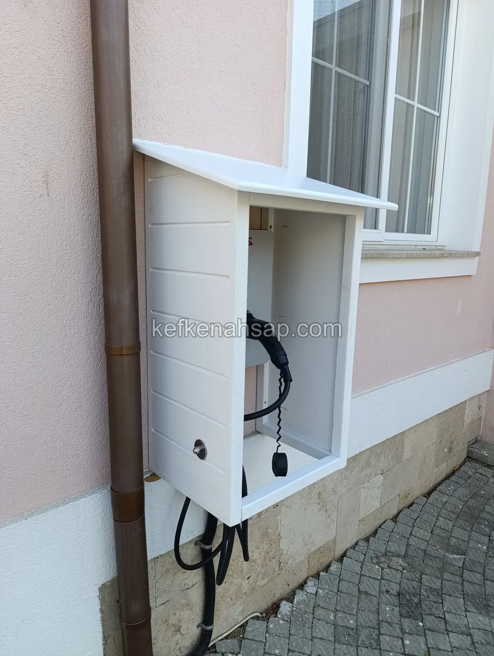 Pembe cephede pencerenin altında yağmur borusunun yanına asılmış, eğimli kapaklı, önü açık beyaz ahşap duvar kabini