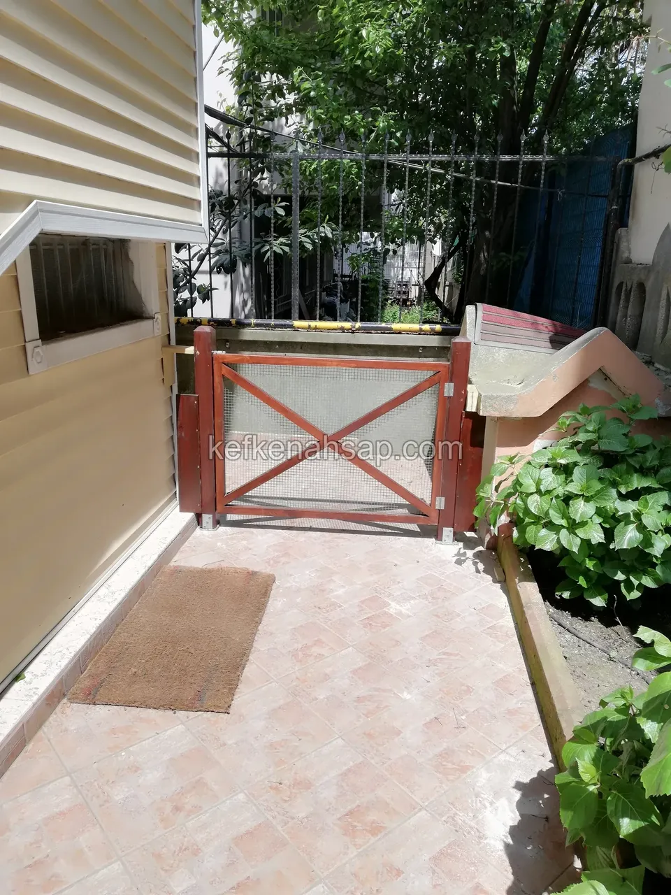 Pembe karo döşeli dar geçitte, X çaprazlı ve tel örgü dolgulu kırmızı boyalı alçak ahşap kapı; solda duvar