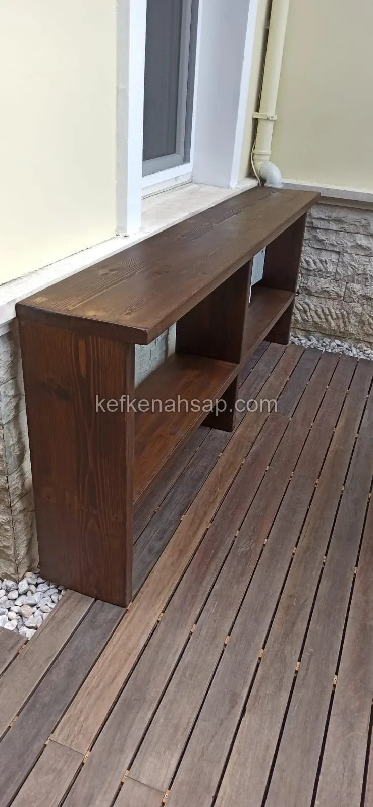 Pencere altında, ahşap deck zemin üzerinde duran koyu kahverengi, uzun ve alçak, açık raflı ahşap ünite