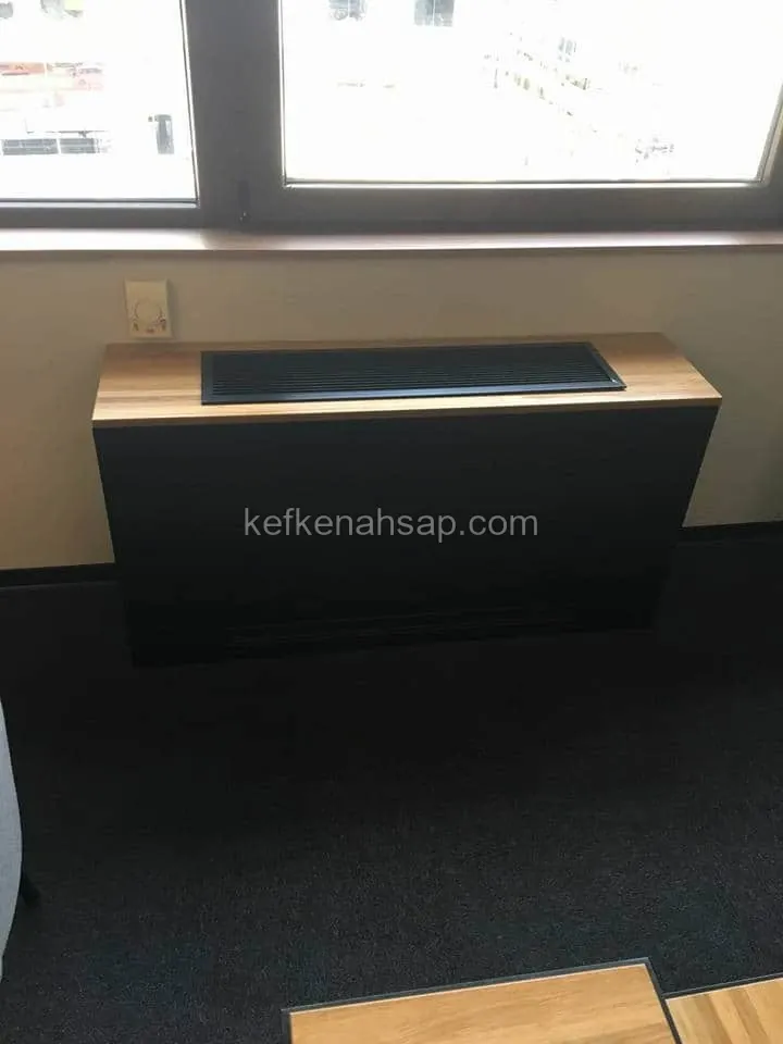 Pencere önünde siyah gövdeli, ahşap üst tablalı ve ortasında siyah ızgara bulunan fan coil kasası