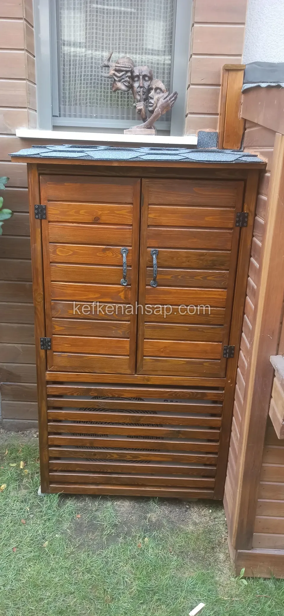 Çimenlik üzerinde, pencerenin altında çift kapaklı ve alt kısmı panjur desenli koyu ahşap dolap