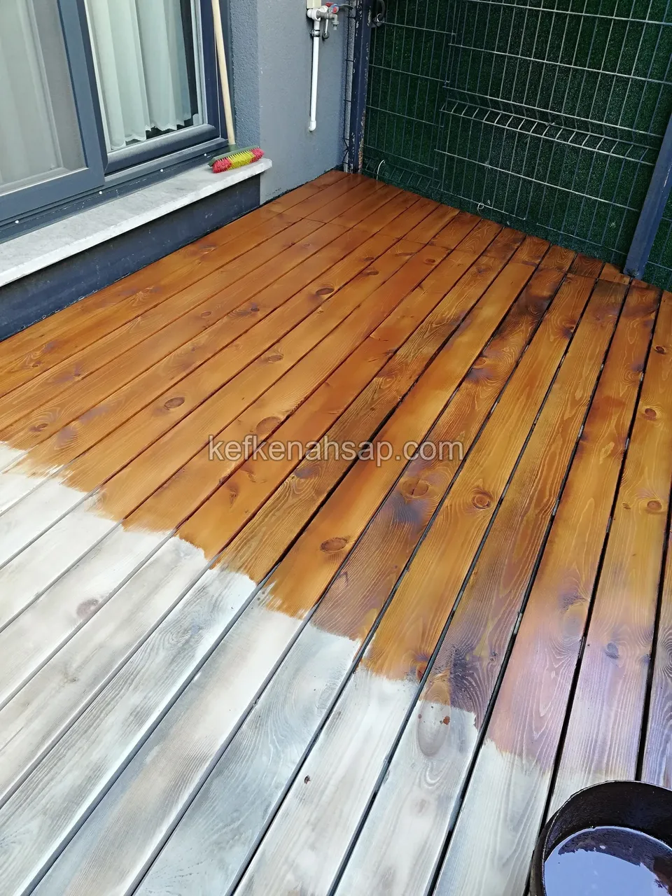 Holzdeck vor einem Fenster, zur Hälfte mit dunklem Holzschutz behandelt, zur Hälfte noch im alten grauen Zustand
