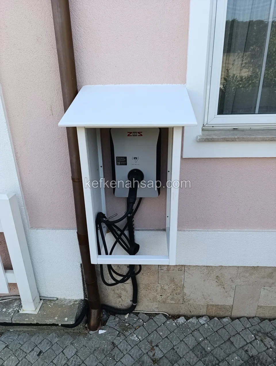 Pembe cephede pencere altına asılmış, eğimli beyaz kapaklı ahşap kabin; içinde duvar cihazı ve kıvrılmış siyah kablo kangalı