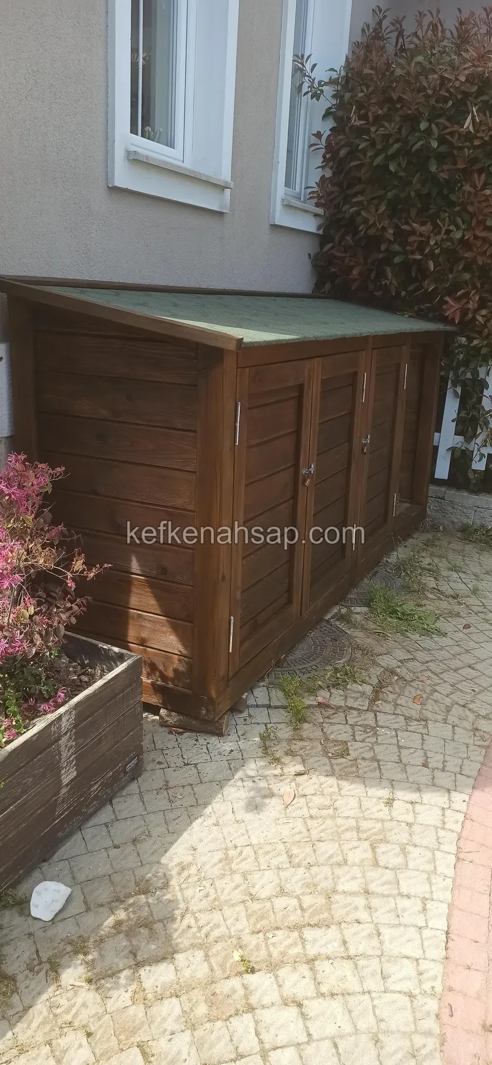 Pencereli duvarın dibinde parke taşlı zeminde yatay tahtalı yüzlü, yeşil kaplı eğimli çatılı uzun alçak ahşap dolap