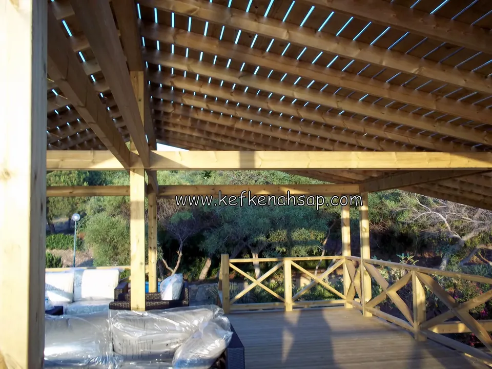 Pergola altında deck, çapraz çıtalı korkuluk ve naylonla sarılı mobilyalar, uzakta ağaçlık yamaç