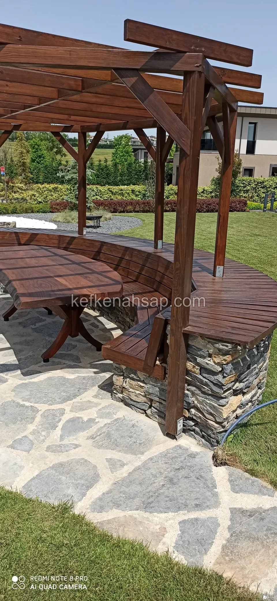 Bahçede pergolanın altında taş kaplı duvarla çevrili kavisli ahşap bank ve ahşap masa, taş döşeli yol
