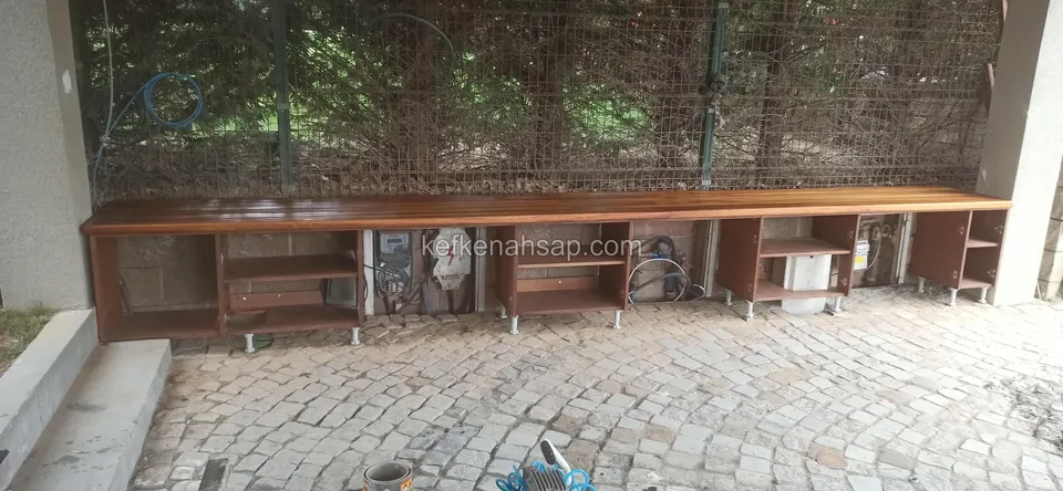Parke taşlı avluda pergola altında duvar boyunca uzanan, geniş tahta tezgâhlı ve açık raflı uzun ahşap dolap hattı