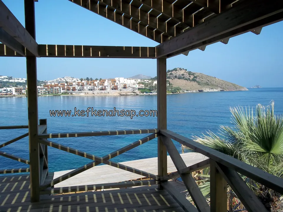 Pergola kirişleri ve çapraz çıtalı korkuluk arasından görünen deniz, palmiye ve kıyıdaki evler