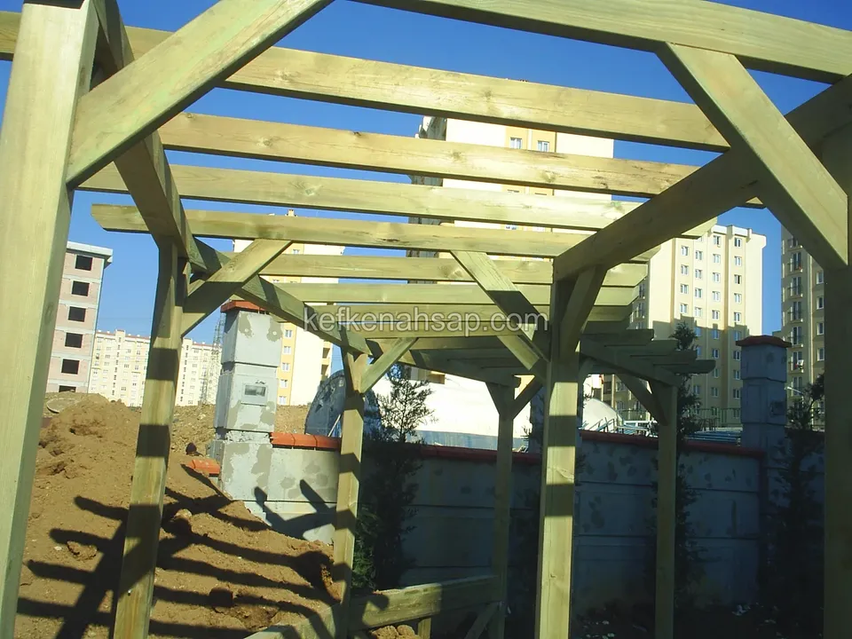 Yakından pergola direkleri, çapraz payandalar ve kirişler, arkada toprak yığını, duvar ve apartmanlar