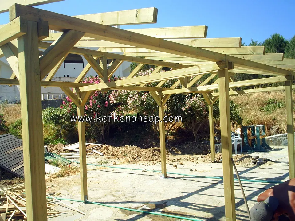 Bahçede uzun pergola direkleri ve üst kirişleri, zeminde kereste artıkları ve çiçekli çalılar