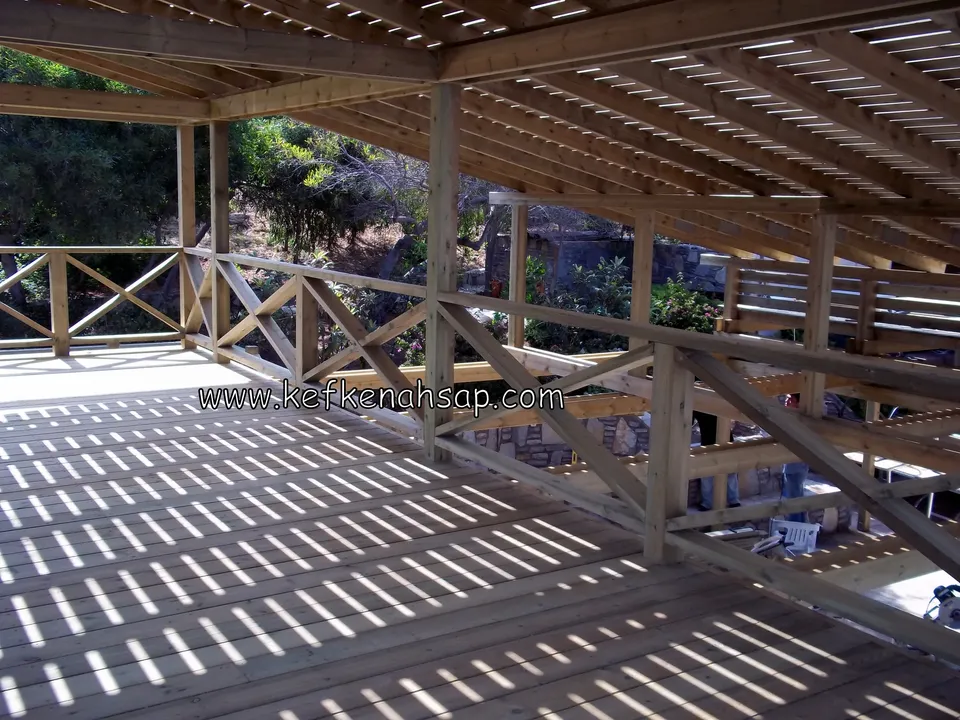 Pergola kirişlerinin gölgelerinin çizgi çizgi düştüğü geniş açık renkli deck zemini ve X korkuluklu kenar