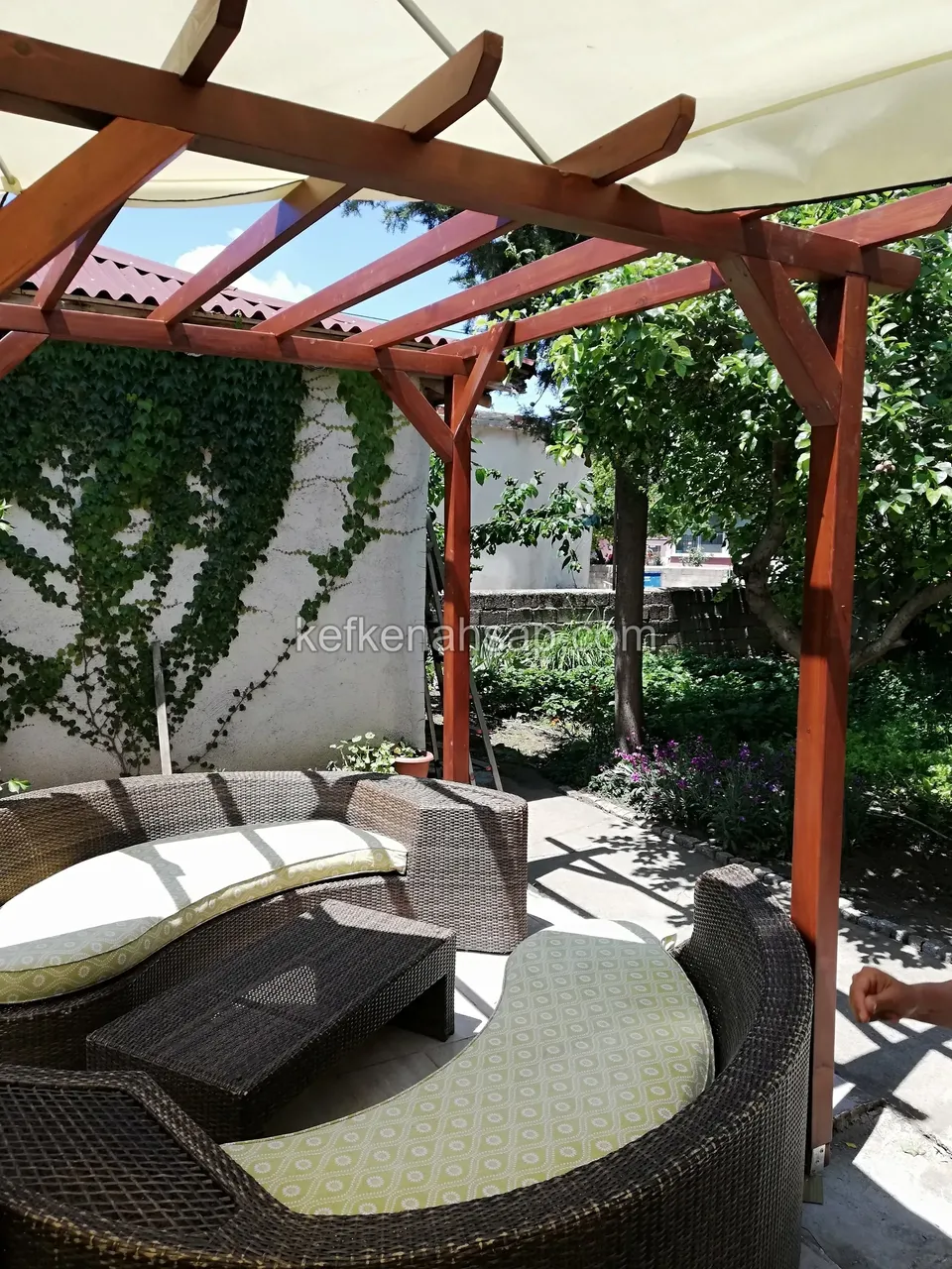 Kırmızımsı pergola kirişlerinin altındaki hasır koltuk takımı ve alçak masa; arkada sarmaşıklı beyaz duvar