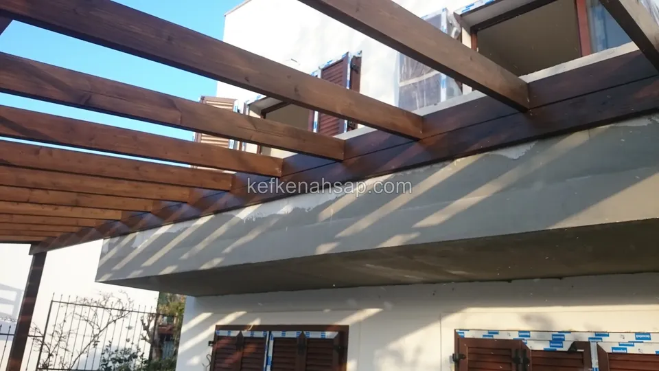 Pergola kirişleri sıvalı balkonun yanından uzanıyor, alt katta kahverengi panjurlu pencereler ve demir korkuluk