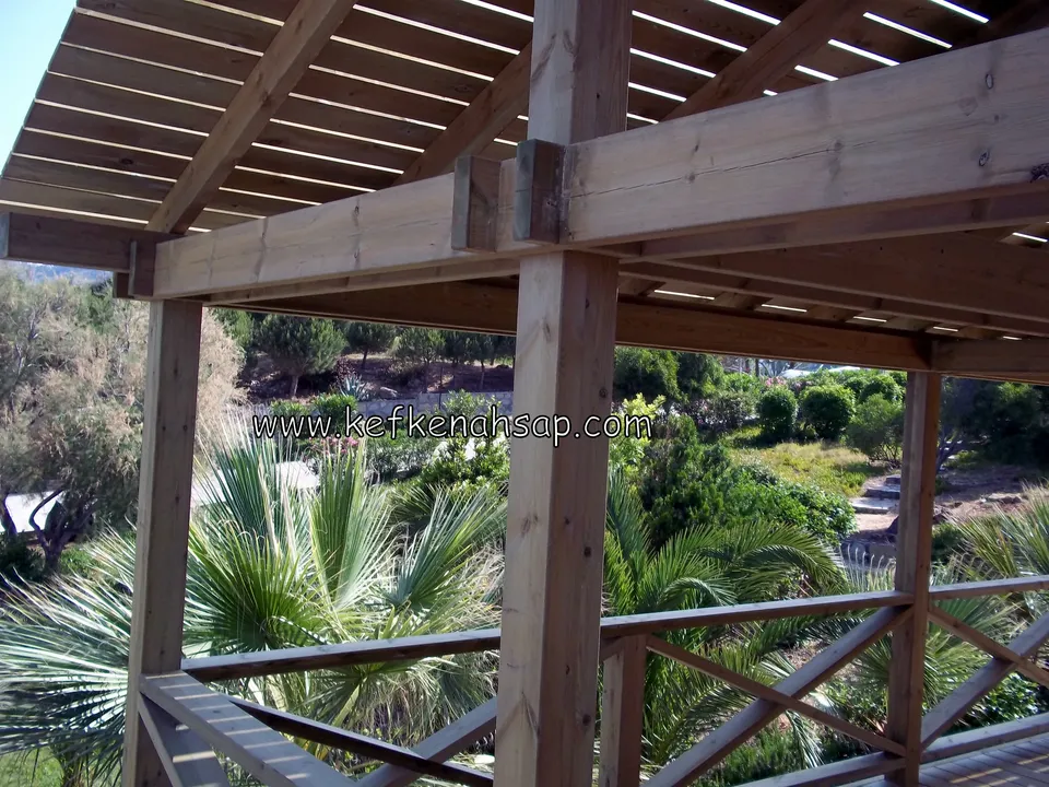 Corner of a pergola where heavy posts and beams meet, with a palm garden beyond cross-braced railings