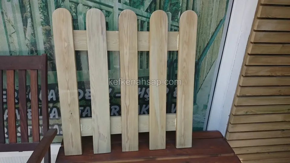 Camlı bir duvarın önünde duran, ucu kavisli dört çıtalı açık tonlu piket çit numune paneli, arkada koyu ahşap sandalye