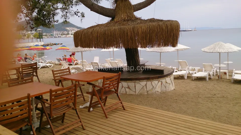 Terrasse um einen Baum mit Strohschirm am Strand, darauf Holztische und Stühle, dahinter Liegen und das Meer