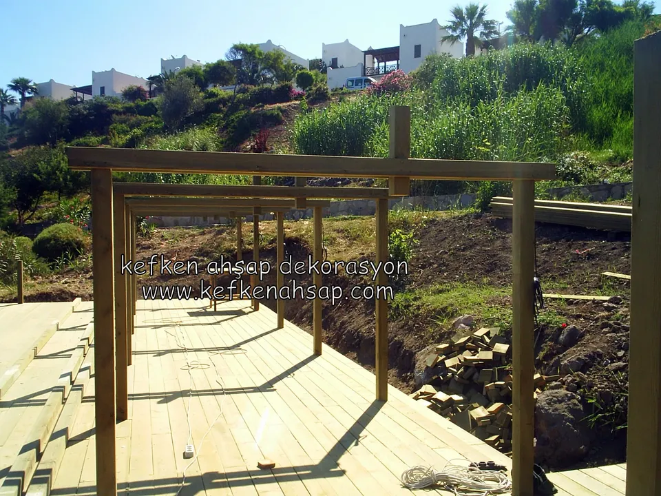 Deck platform üzerinde yan yana sıralanan kalın ahşap direkler ve üstlerinde yatay kirişler, arkada yamaç ve beyaz evler