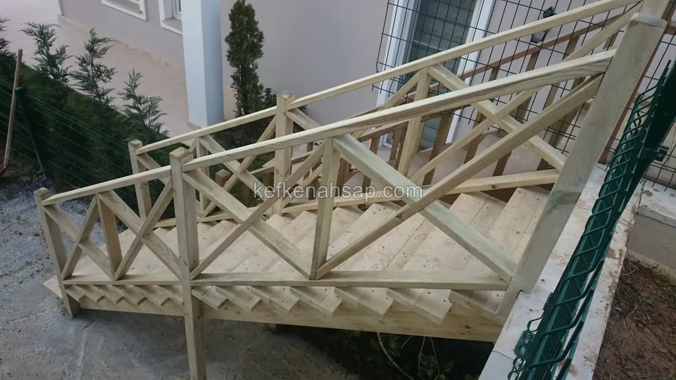 Yandan görünen platformlu ahşap merdiven; çaprazlı korkuluk, yanda yeşil telli çit ve ev cephesi