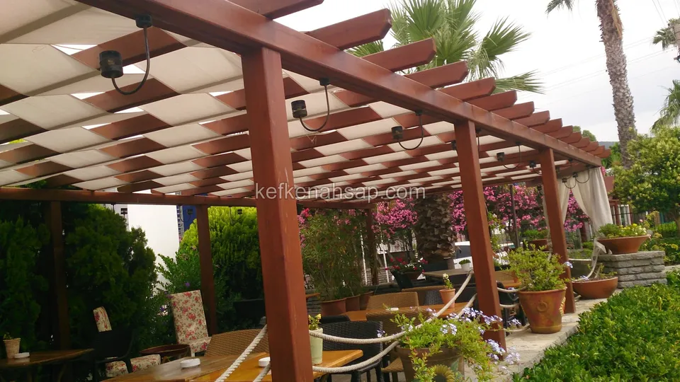 برجولا خشبية مطلية بالورنيش المائل للحمرة في حديقة مطعم بغطاء قماشي كريمي وأصص زهور ونخيل