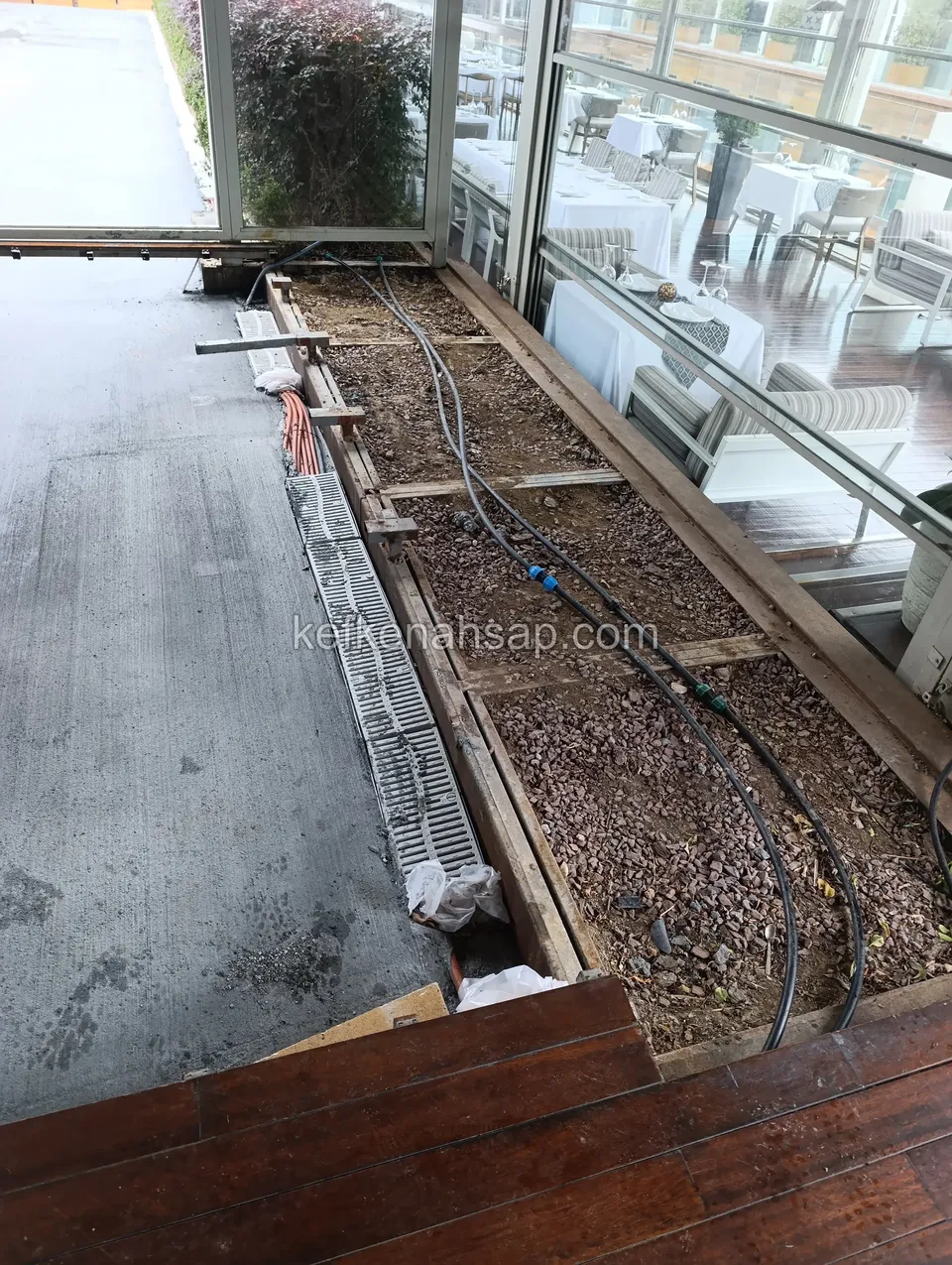 Cam cepheli restoranın önünde, deck çıkarılmış alanda açıkta kalan çakıl, borular ve su ızgarası; altta deck tahtaları