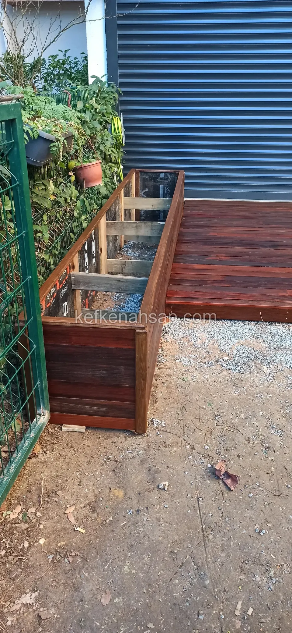 Dunkler Holzpflanzkasten neben einem Rollladen, daneben ein rotbraunes Deck