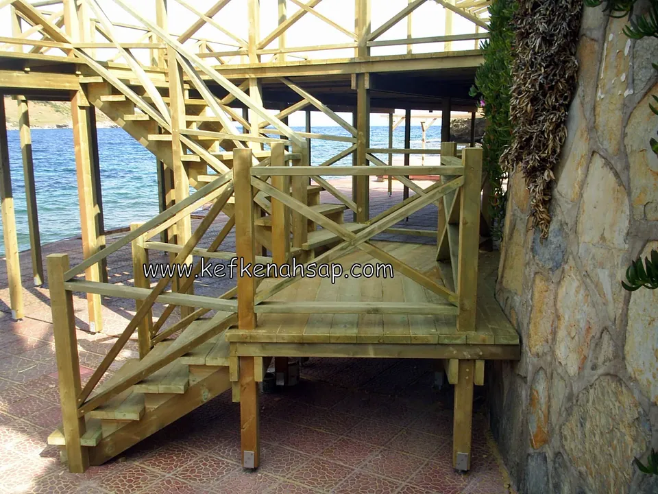 Holztreppe mit Diagonalgeländer, die an einer Steinmauer vom Podest nach oben abknickt, dahinter das Meer