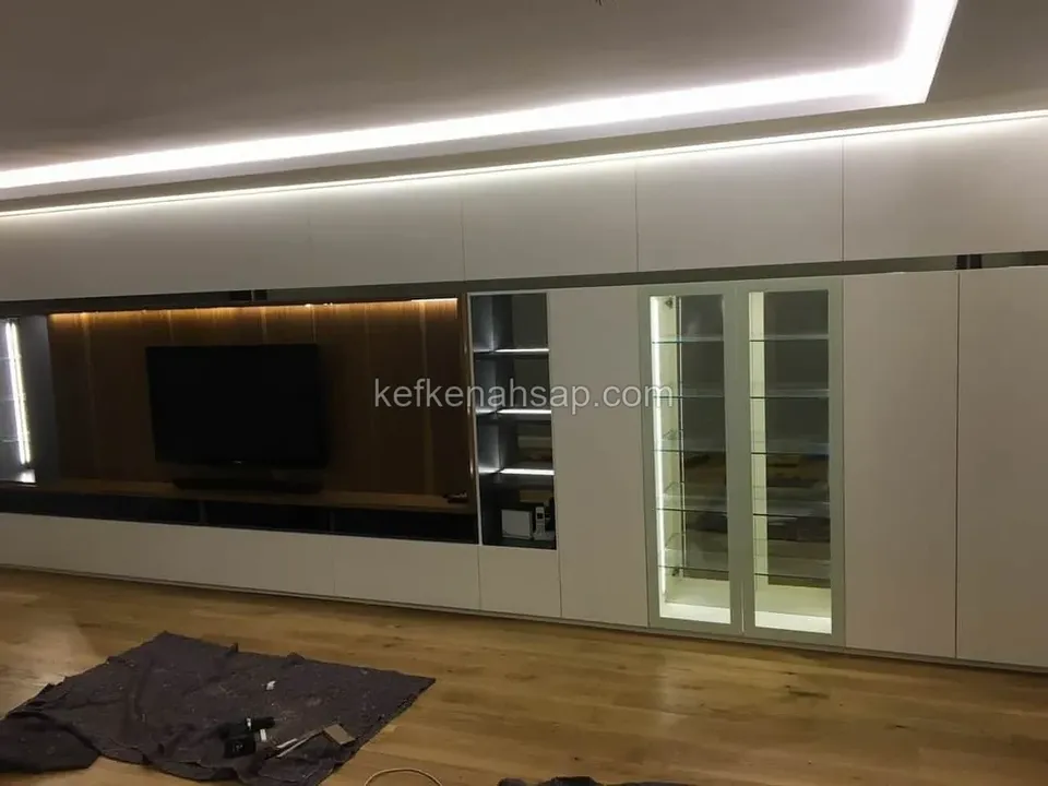 Salonda beyaz mat dolaplar, ahşap kaplı TV bölümü, camlı ışıklı vitrin ve tavanda gizli LED aydınlatma