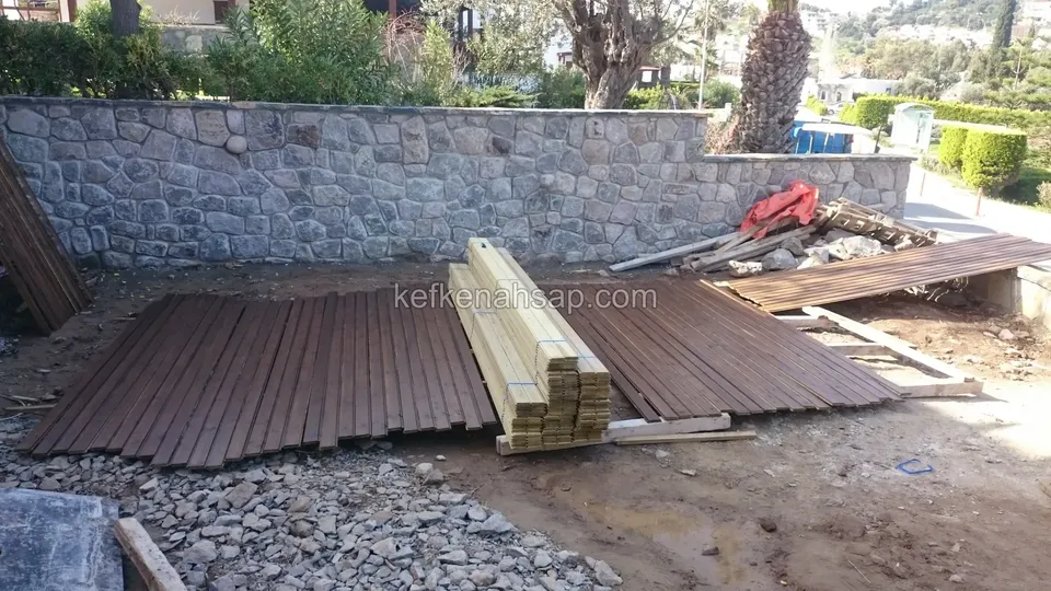 Çakıllı zeminde yerde duran koyu deck tahtaları, bağlı kereste destesi ve taş duvar