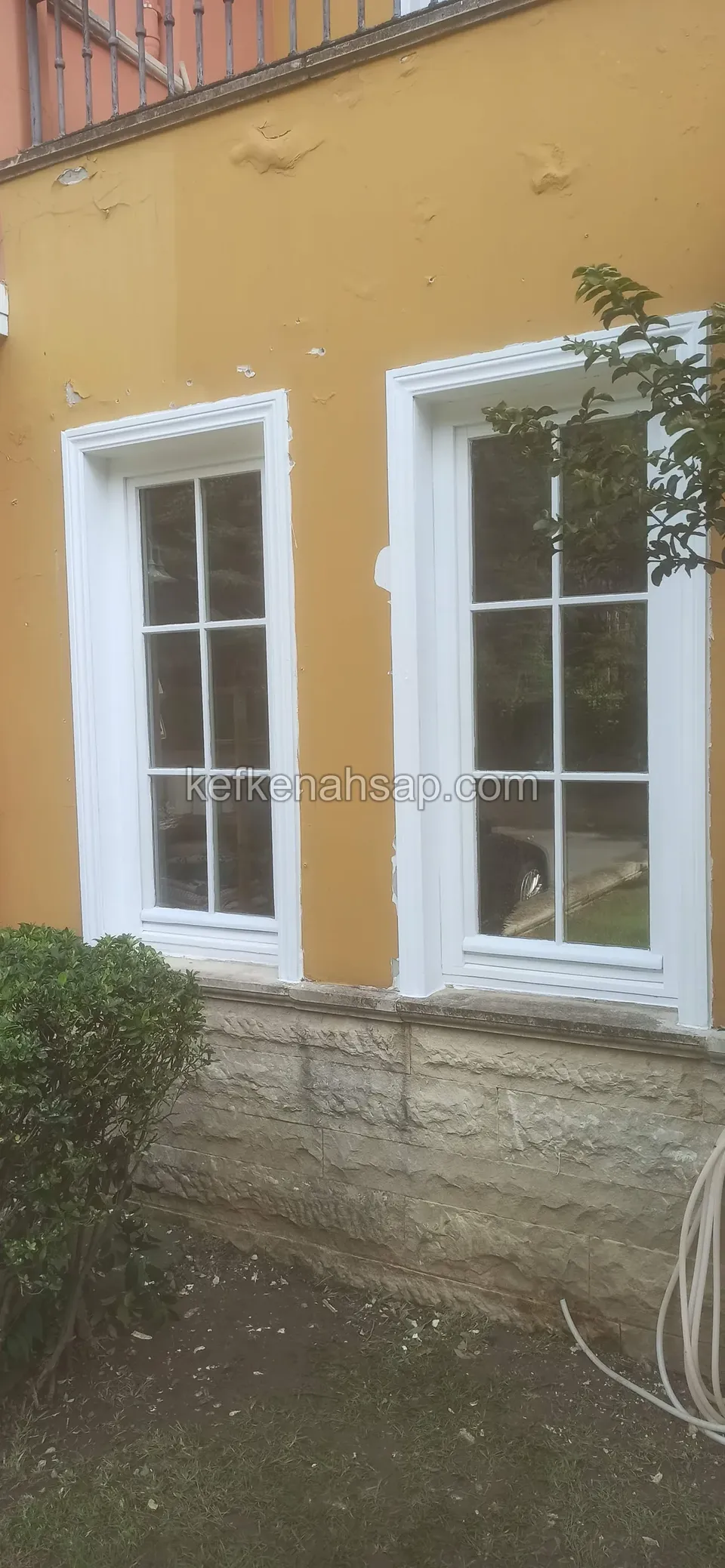 Sarı boyalı cephede, taş kaplı lambri üzerinde duran beyaz ahşap doğramalı iki pencere, kare cam bölmeli