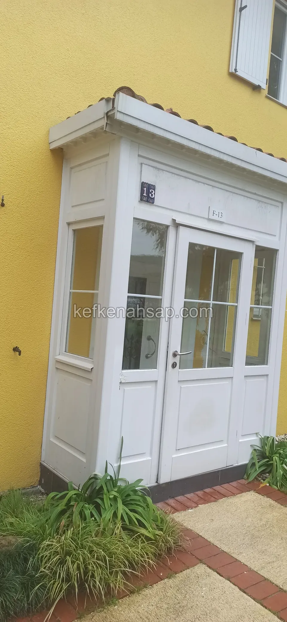 Sarı boyalı bir evin girişinde, beyaz ahşap doğramalı ve camlı küçük bir giriş kabini, önünde kırmızı tuğla kenarlı yol
