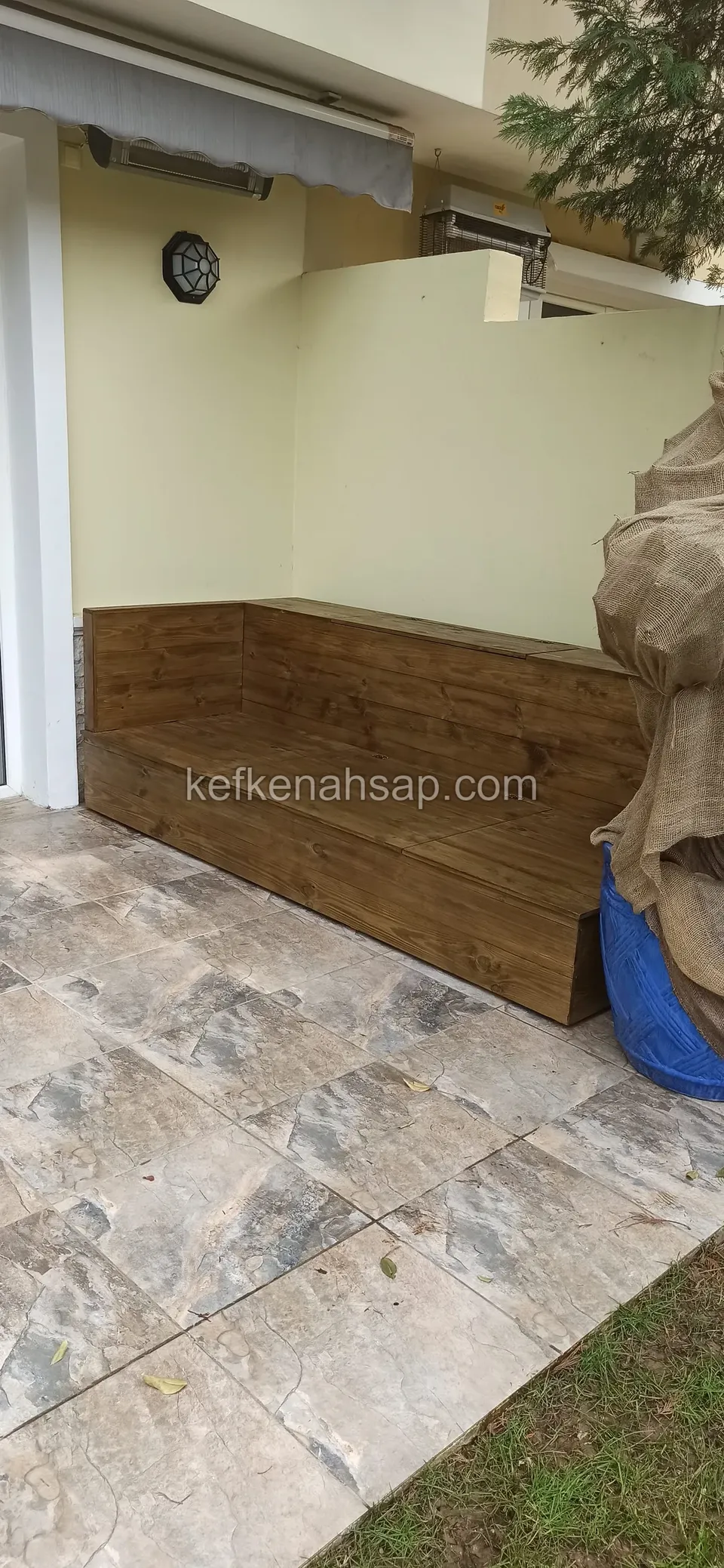 Eine dunkel lackierte Holz-Eckbank, in die Ecke gelb gestrichener Terrassenwände eingepasst, davor Fliesen in Steinoptik