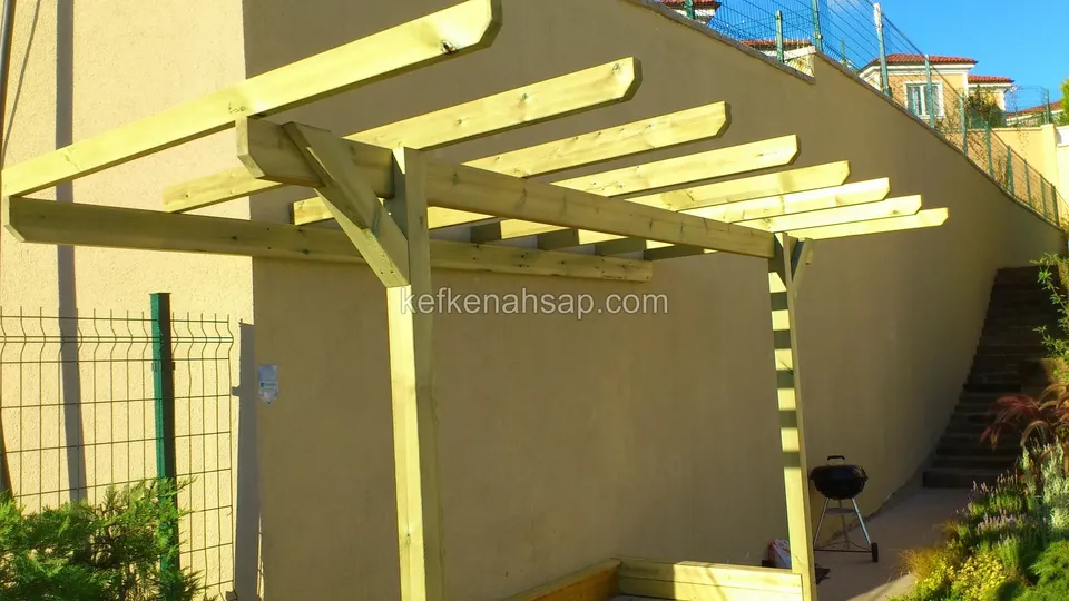 Sarı sıvalı yüksek duvara yaslanan, açık renk ahşaptan tek sıra dikmeli pergola
